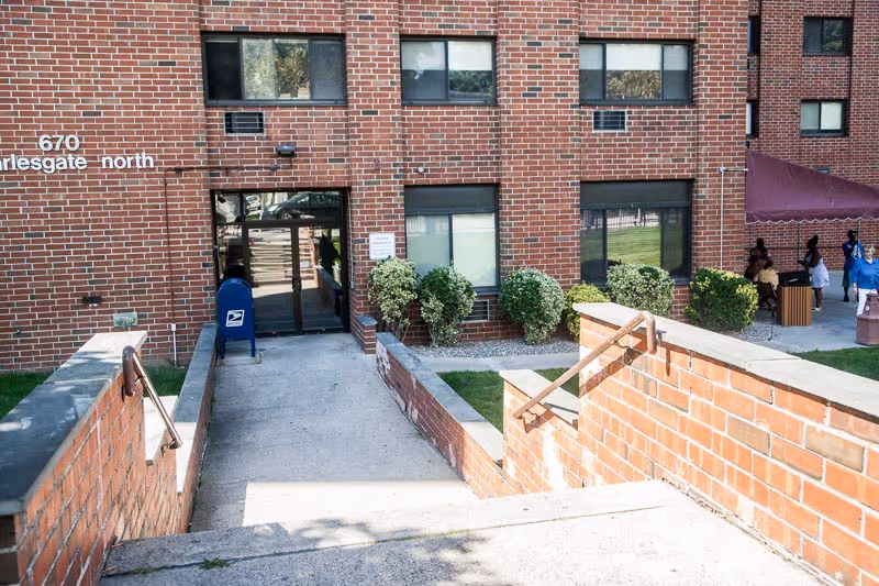 Brick-faced nursing center entrance with concrete ramp and stairs, a blue USPS mailbox, and people seated near the building.