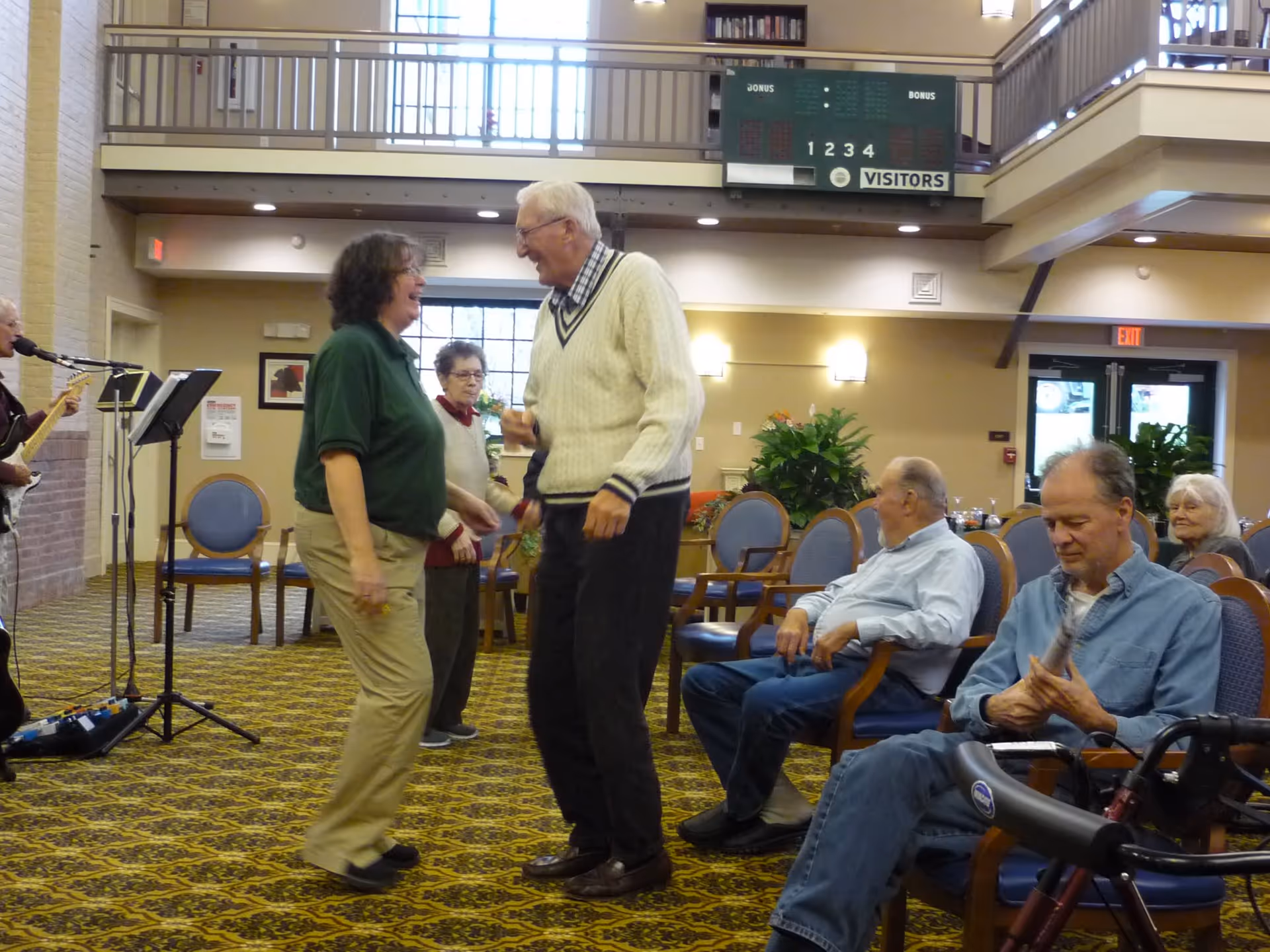 A group of elderly people and a staff member enjoying a social activity in a senior community common area. Two people are standing and smiling at each other in the center, while others are seated on chairs around them. A musician is playing guitar and singing on the left side. The room has patterned carpet, chairs with blue cushions, and a scoreboard on the wall above.