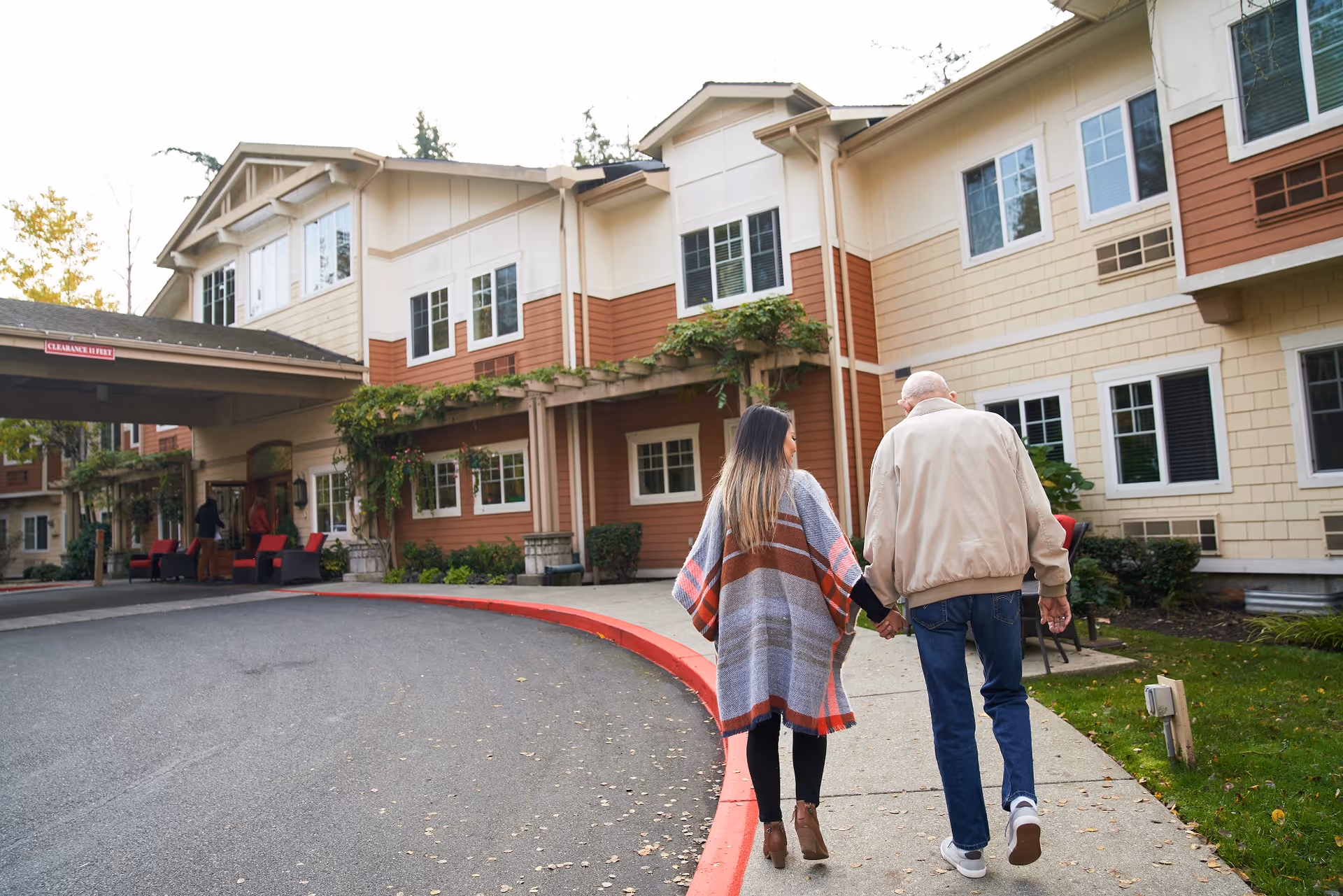 A woman and an elderly man walking hand-in-hand toward the entrance of a multi-story senior living building.