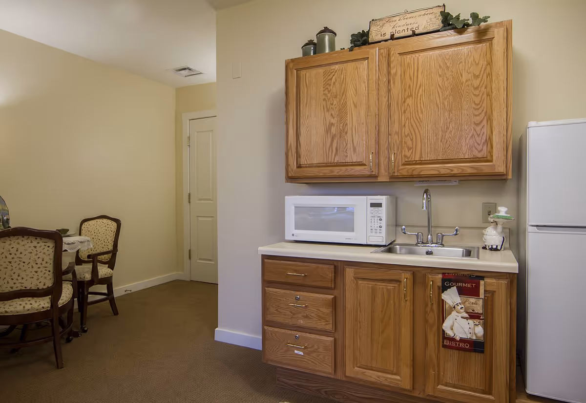 A small kitchen area with wooden cabinets, a white microwave on the countertop, a stainless steel sink with a faucet, and a white refrigerator. To the left, there is a dining area with a round table and four upholstered chairs with wooden frames. The walls are painted beige and the floor is carpeted.