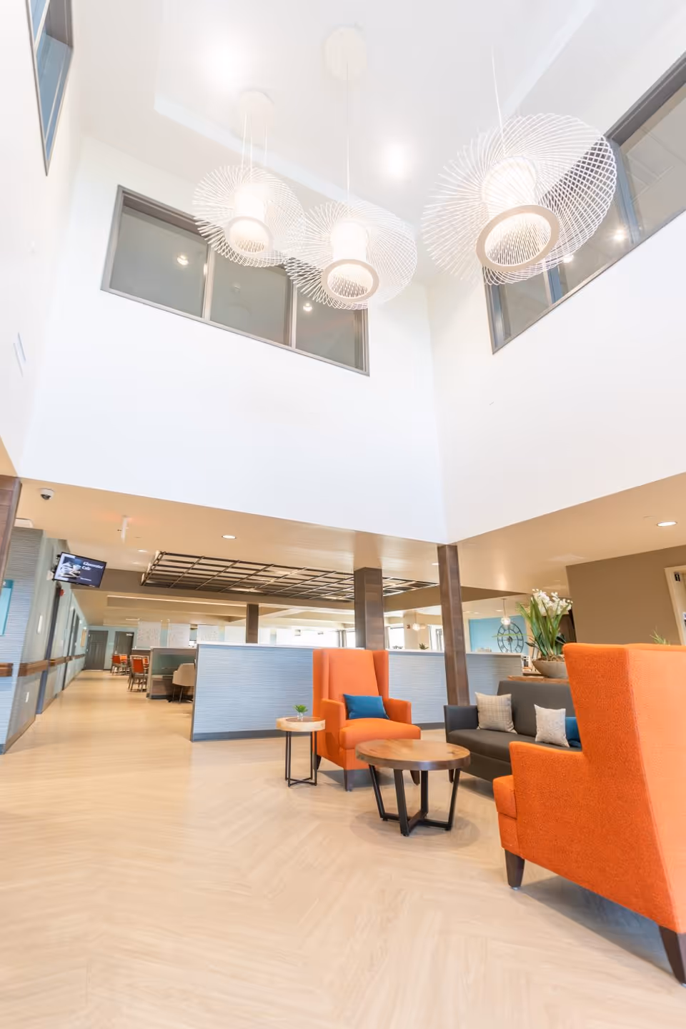 Bright and spacious senior living facility common area with high ceilings, modern hanging light fixtures, orange armchairs with blue cushions, a gray sofa with pillows, and round wooden tables. The area has light wood flooring and large windows on the upper walls.