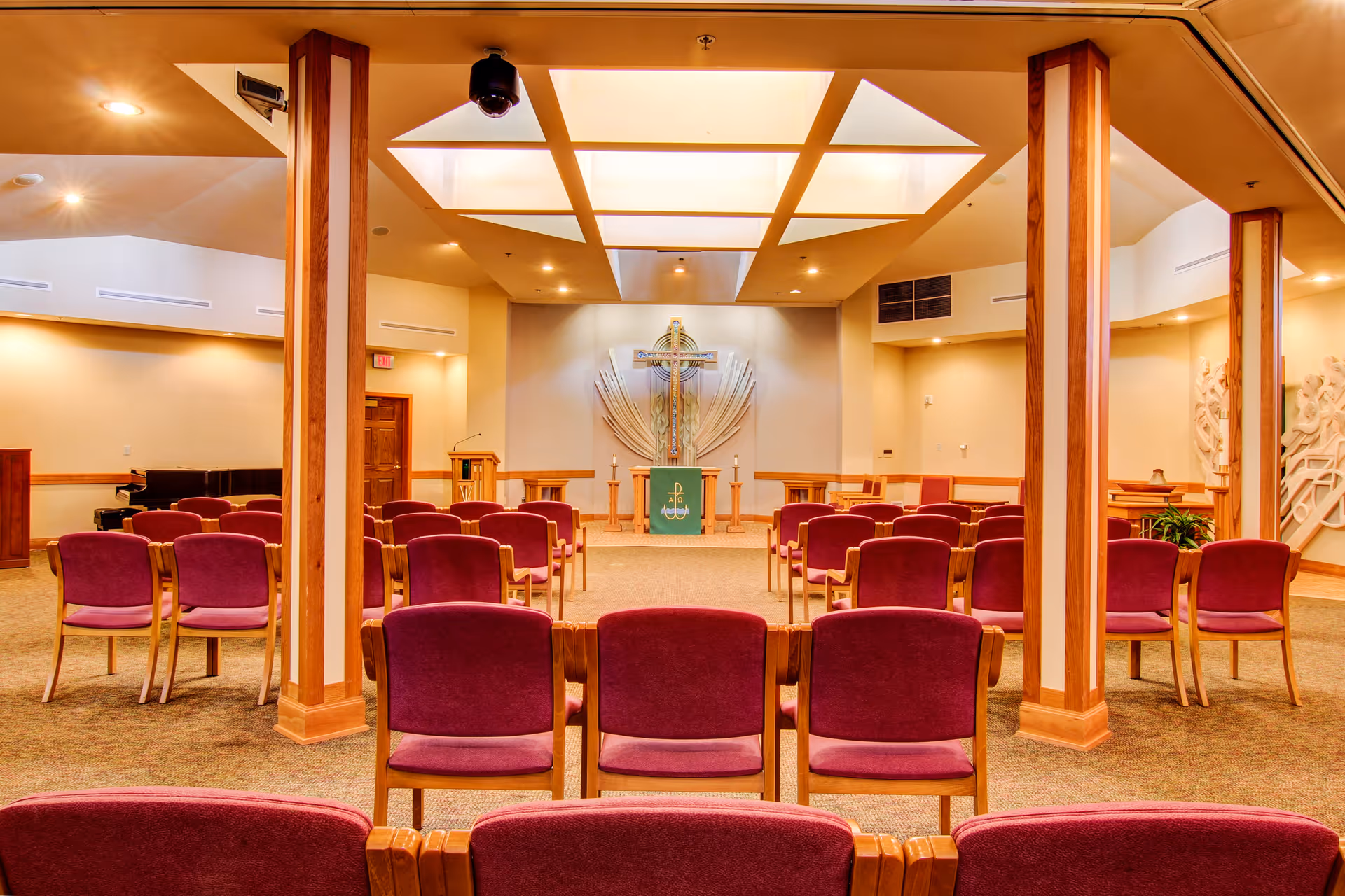 Interior view of a chapel or worship room with rows of purple cushioned chairs facing a wooden altar adorned with a green cloth and a large cross on the wall behind it. The room has warm lighting, wooden columns, a pulpit, and a piano in the corner.