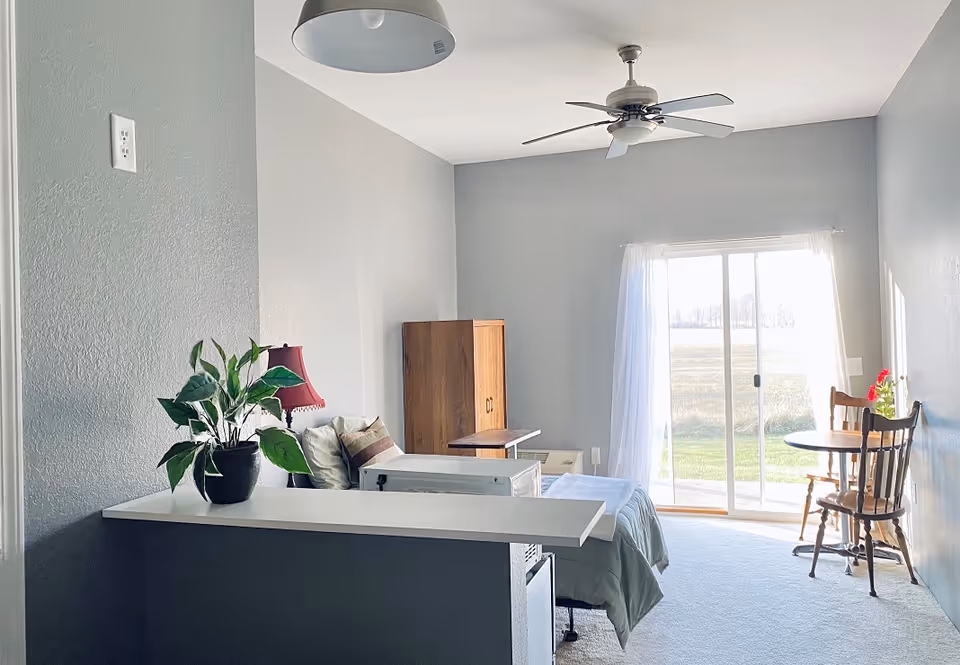 Bright studio bedroom with a bed, dresser, ceiling fan, potted plant on a counter and a small table and chairs by a sliding glass door.