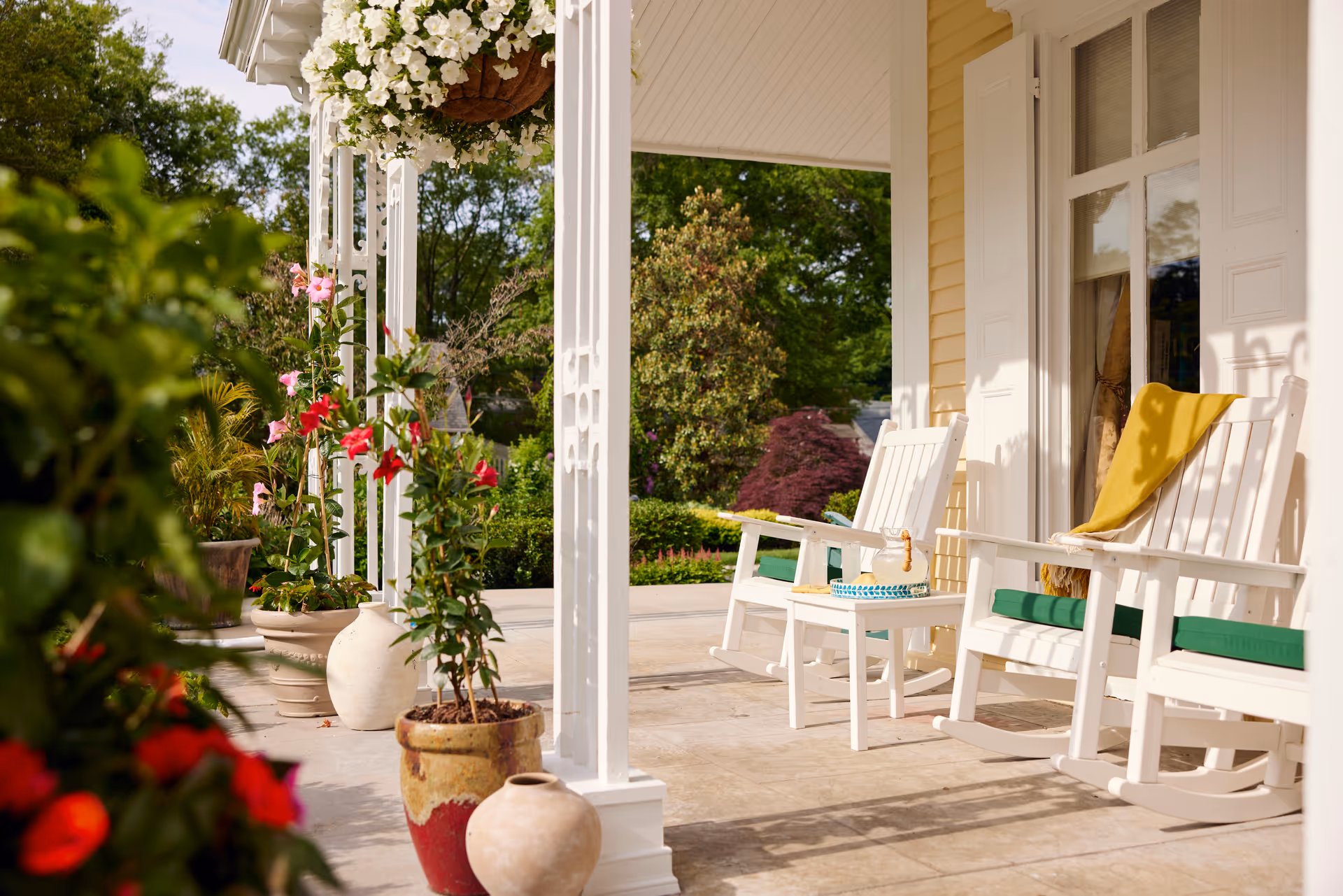 A sunny porch area with white rocking chairs, green cushions, and a yellow blanket draped over one chair. There are several potted plants with colorful flowers and greenery around the porch, and a hanging basket with white flowers. The porch is attached to a light yellow building with white trim and shutters.