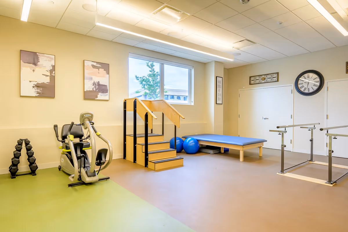 A bright physical therapy or exercise room with light-colored walls and ceiling. The room features exercise equipment including a recumbent exercise bike, a set of dumbbells, a wooden staircase with handrails, two blue exercise balls, a padded therapy table, and parallel bars for walking exercises. There are two abstract paintings on the wall and a large clock above two closed white doors.