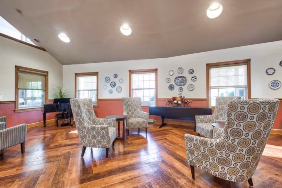 A bright and spacious common area with wooden herringbone flooring, four patterned armchairs arranged around a small round table, a black grand piano in the corner, three windows with wooden frames, and decorative blue and white plates mounted on the walls.