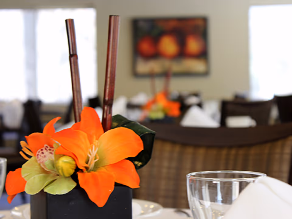 Close-up of a dining table centerpiece featuring bright orange flowers and two tall brown sticks in a black square vase, with blurred tables and chairs in the background in a dining room setting.