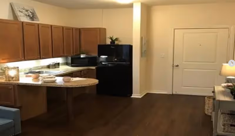 Interior view of a kitchen area with wooden cabinets, a black refrigerator, a microwave, and a countertop with two cups and plates. The floor is dark wood, and there is a white door and a small table with a lamp on the right side.