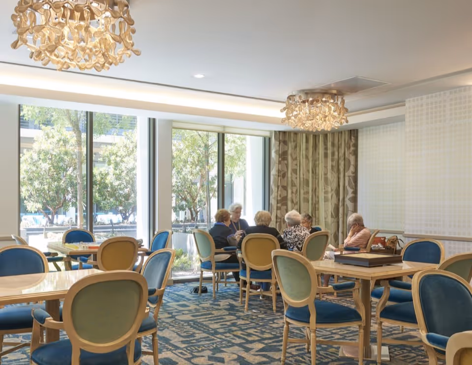 Bright communal dining/activity room with round tables and blue-upholstered chairs and several seniors seated near large windows.