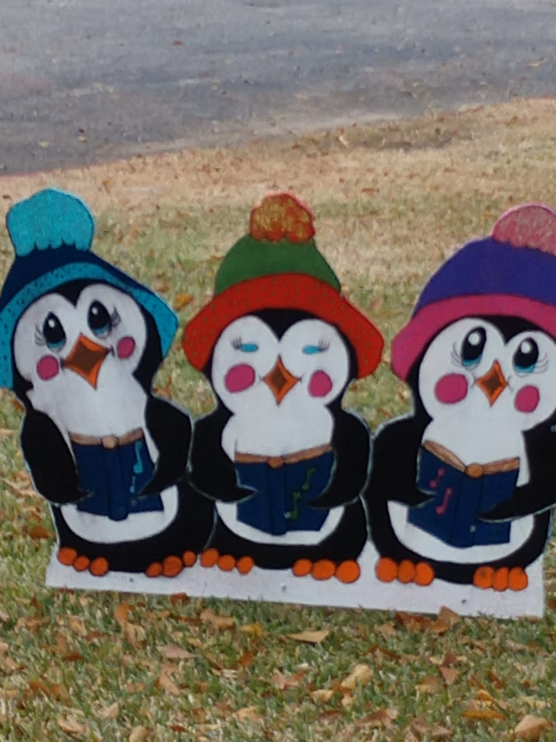 A decorative outdoor display featuring three cartoon penguins wearing colorful winter hats and holding blue songbooks, standing on grass with a paved area in the background.