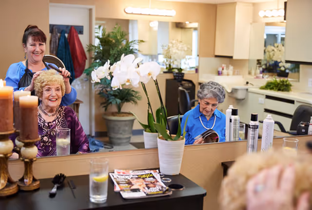 Two elderly women in a salon area—one getting her hair styled while another reads a magazine—reflected in a mirror with plants and salon products on the counter.