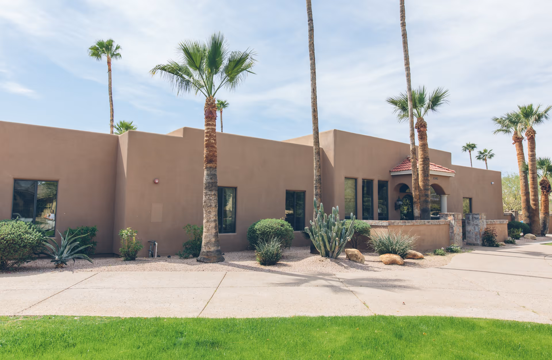 Exterior view of a single-story building with a southwestern architectural style, featuring tan stucco walls, several windows, and a small entrance with a red-tiled roof. The building is surrounded by desert landscaping including palm trees, cacti, and shrubs, with a green lawn and a paved walkway in the foreground under a partly cloudy sky.