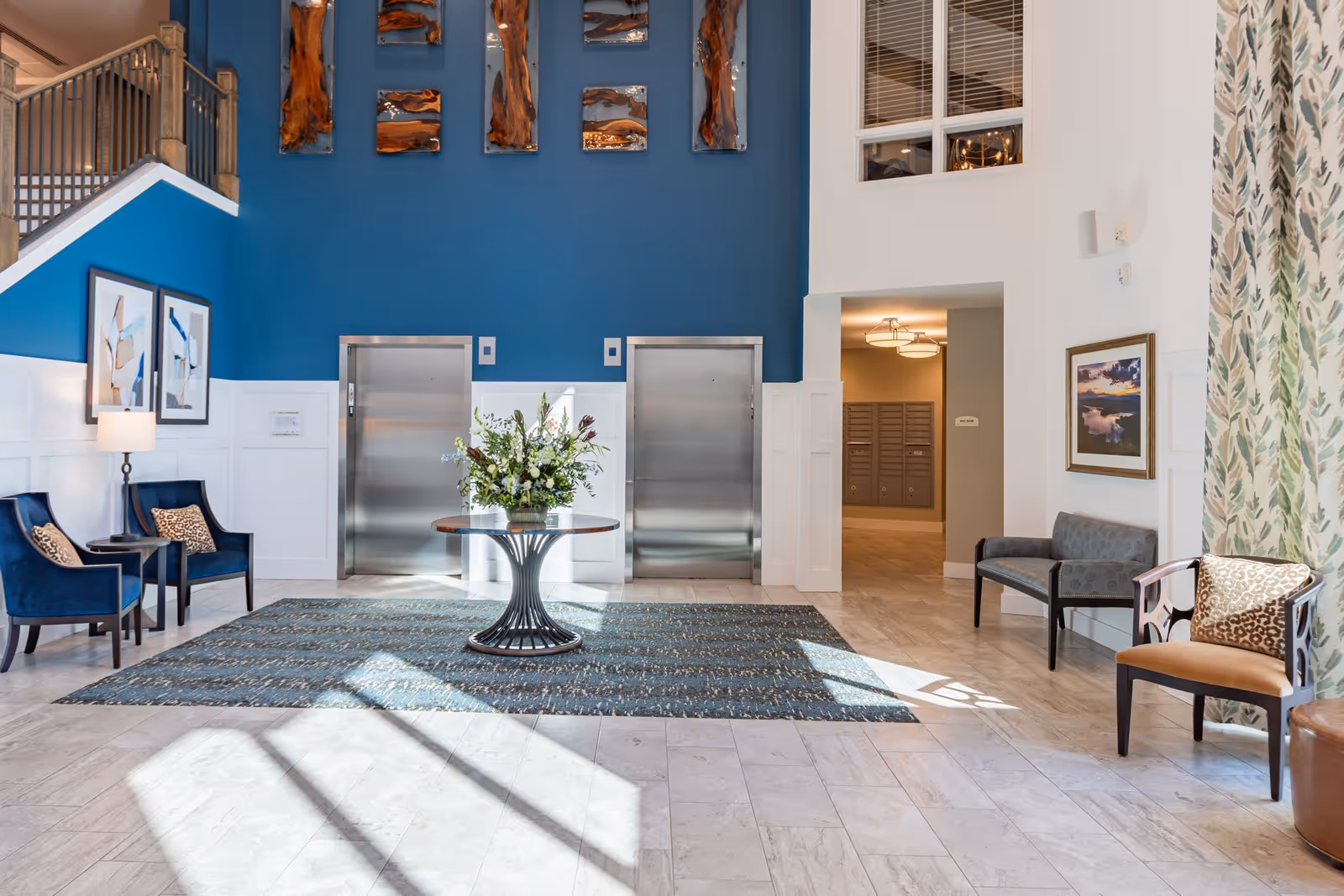 A bright and spacious lobby area with two stainless steel elevators set into a blue accent wall. In front of the elevators is a round table with a large floral arrangement. The floor is tiled with a large patterned rug in the center. To the left are two blue upholstered chairs with leopard print pillows and a small side table with a lamp. To the right are additional seating options including a bench and a chair with a leopard print pillow. The walls are decorated with framed artwork and wood art pieces above the elevators. Sunlight streams in through large windows, casting shadows on the floor.