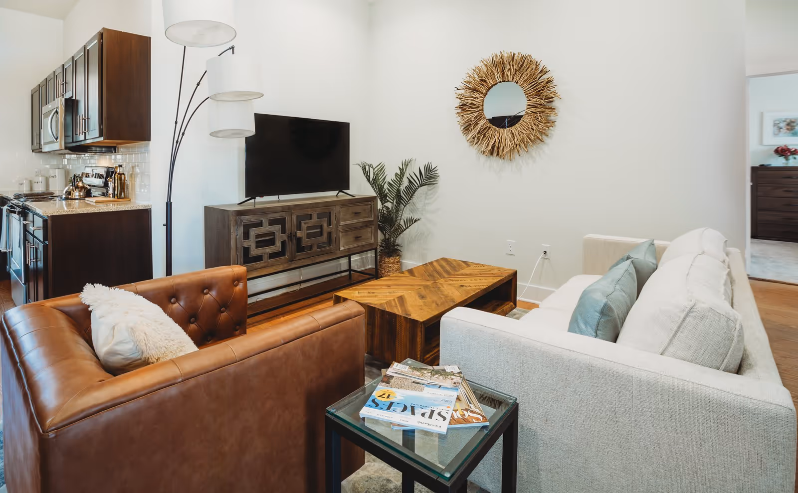 A cozy living room area with a white sofa and a brown leather armchair, a wooden coffee table, a glass side table with magazines, a TV on a wooden console, a tall floor lamp, a decorative round mirror with a natural frame on the wall, and a potted plant in the corner. The kitchen with dark wooden cabinets and stainless steel appliances is visible to the left.
