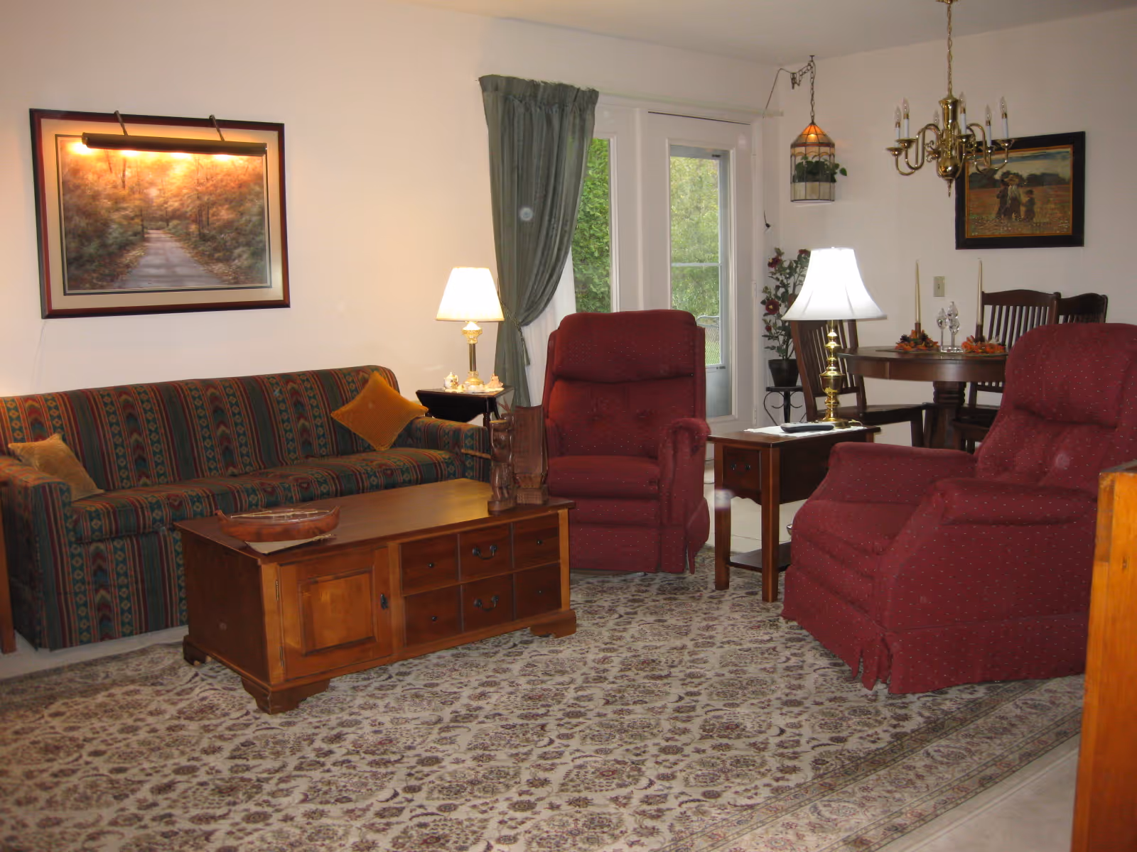Cozy living room with a patterned sofa, two red recliners, a wooden coffee table, lamps, and a dining table near glass doors.