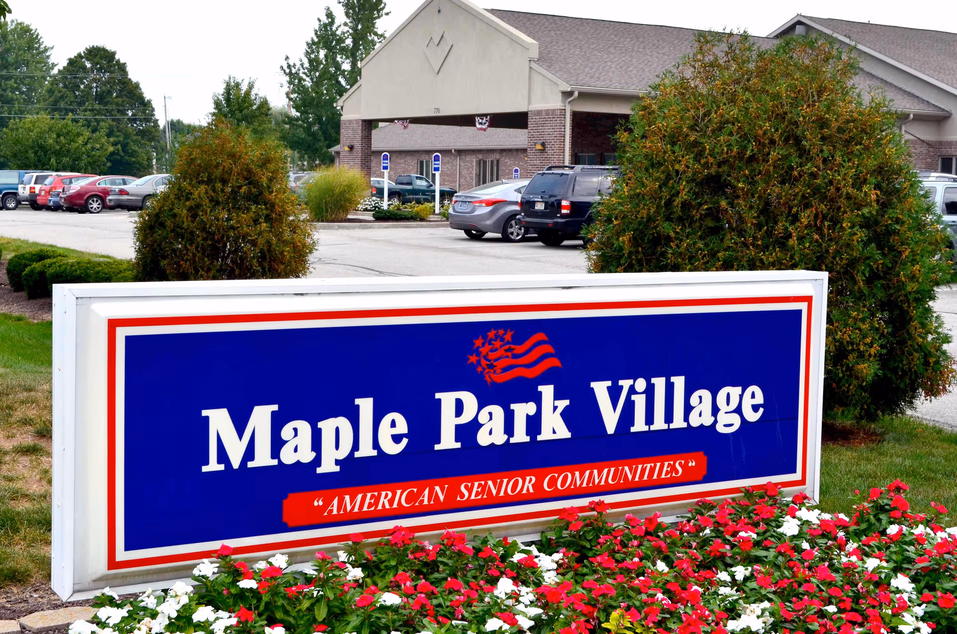 Outdoor view of the entrance area of Maple Park Village senior living community, featuring a large blue sign with white and red text that reads 'Maple Park Village' and 'American Senior Communities'. The sign is surrounded by red and white flowers, with bushes and parked cars visible in the background near a brick building.