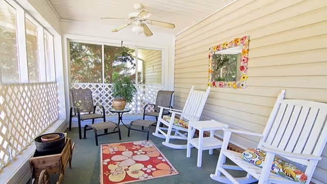 A screened-in porch with two white rocking chairs with floral cushions, two black chairs, a small round glass table with a potted plant, a colorful floral rug on the floor, and a ceiling fan above. The walls are beige with horizontal siding and a decorative mirror with a flower frame hangs on one wall.