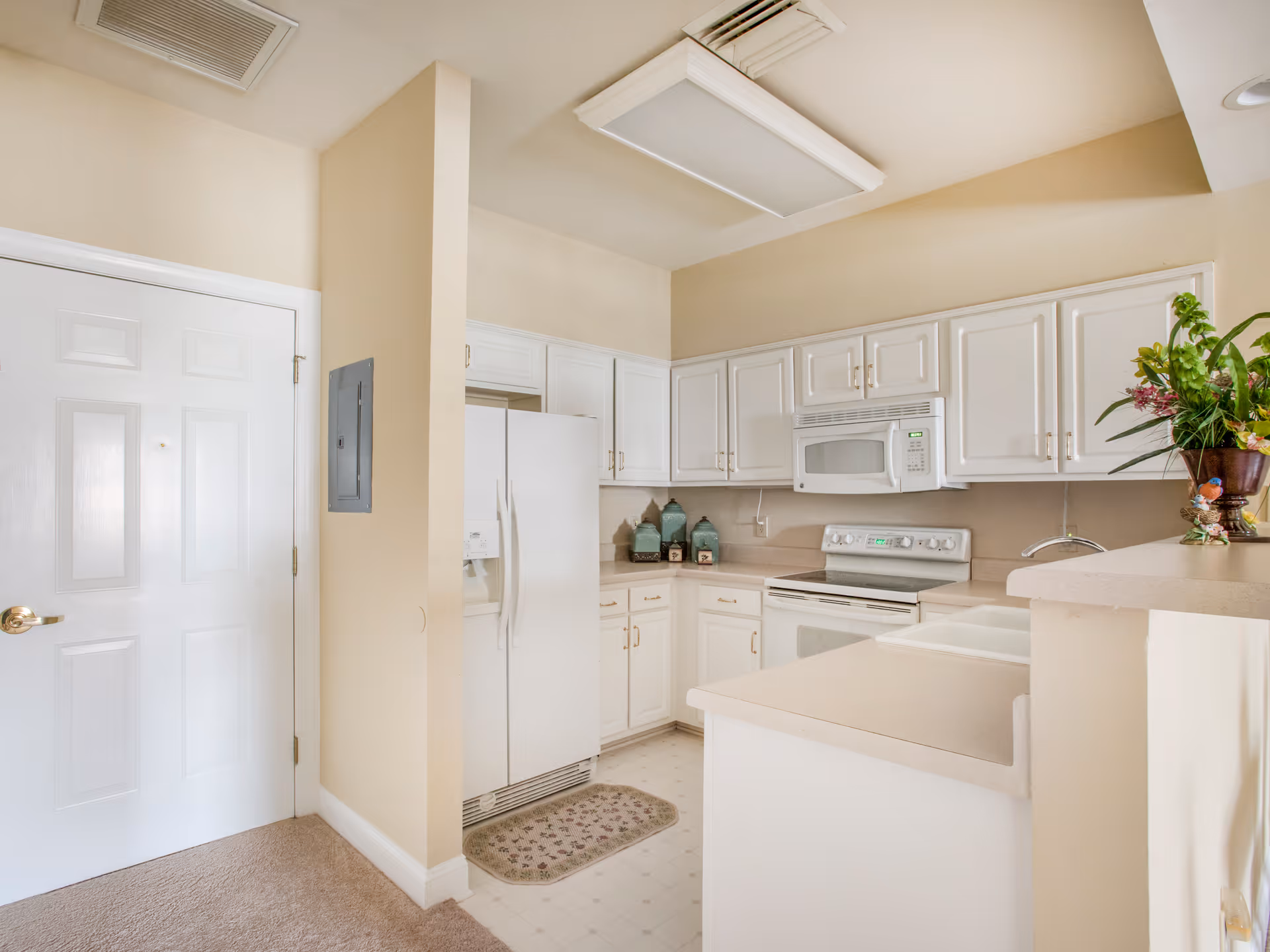 Bright kitchen with white cabinets, a white refrigerator, stove, and microwave. There is a beige countertop with a sink and a decorative plant on the right side. The walls are painted a light beige color, and there is a white door on the left side of the image.