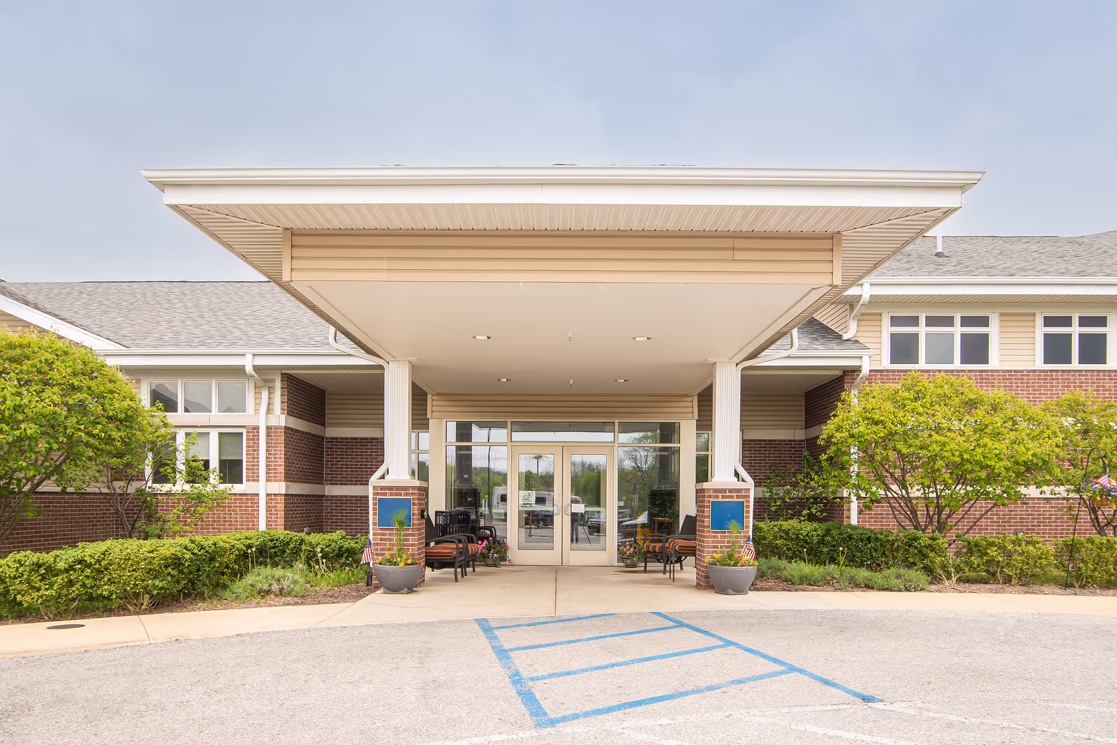 Front entrance of a senior living facility with a covered drop-off area, glass double doors, brick and beige siding exterior, and landscaped greenery on both sides.