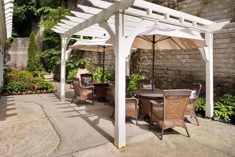Outdoor patio area with a white pergola providing shade over two round tables with wicker chairs and beige umbrellas. The patio is surrounded by a stone wall and greenery, including potted plants and bushes.