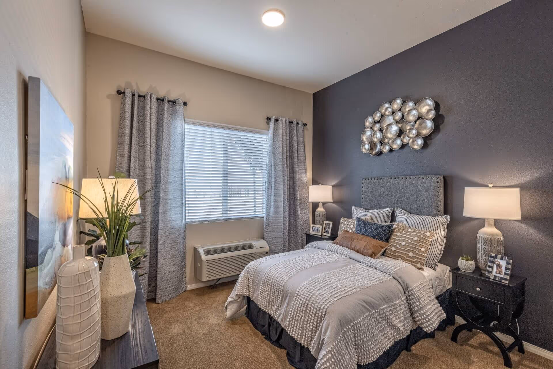 A cozy bedroom with a bed featuring multiple pillows and a textured gray comforter. The room has a dark accent wall behind the bed with a decorative metallic wall art piece. There are two bedside tables with lamps and framed photos. A window with gray curtains and blinds lets in natural light. A dresser with decorative vases and a lamp is positioned opposite the bed.