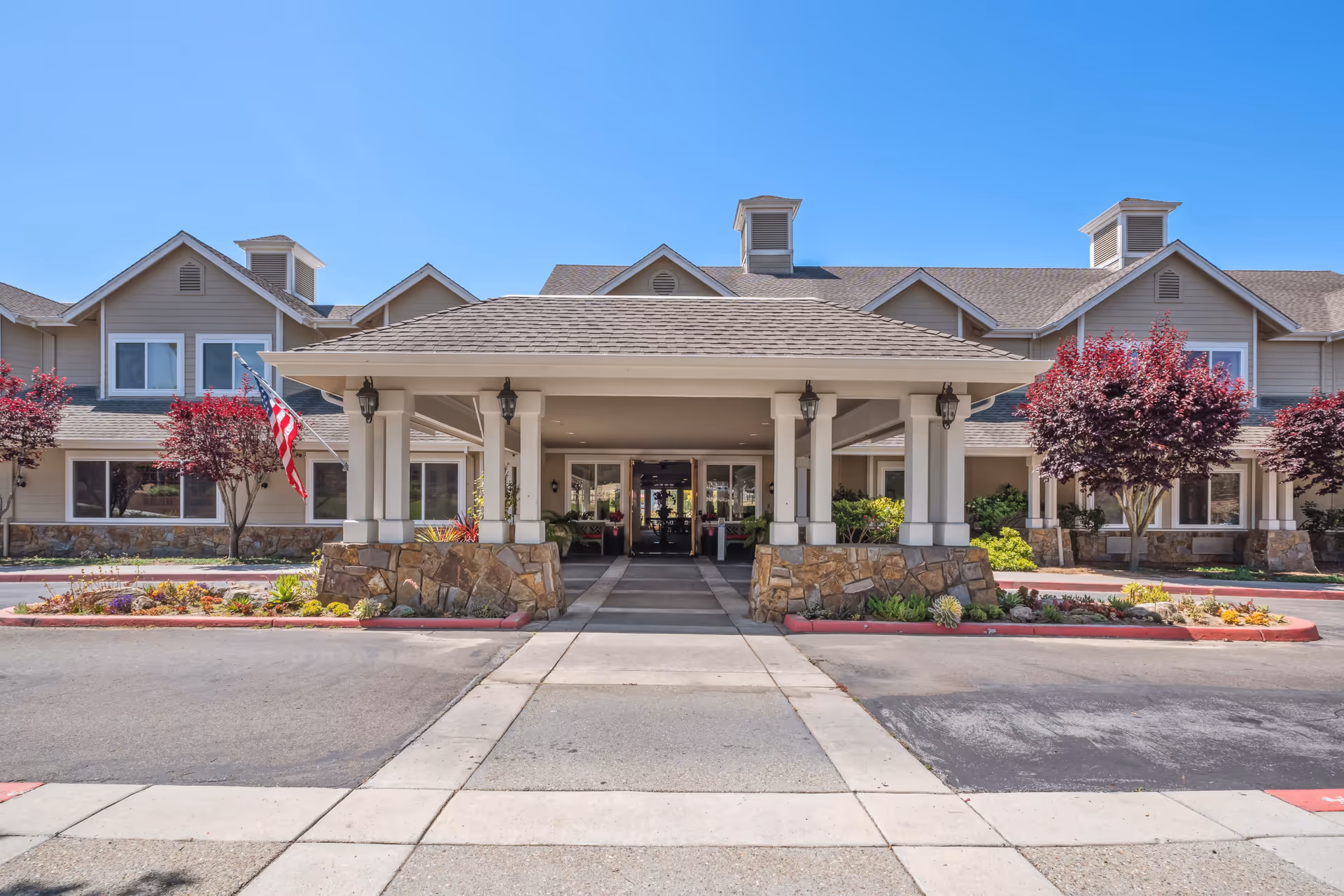 Front exterior view of Aegis Living Aptos facility with a covered entrance, stone pillars, landscaped flower beds, and trees with red foliage under a clear blue sky.