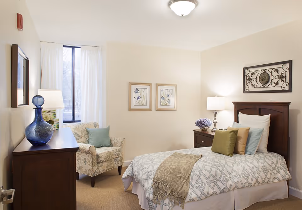 A cozy bedroom with a single bed adorned with multiple pillows and a patterned blanket. Next to the bed is a wooden nightstand with a lamp and a small vase of flowers. There is a patterned armchair with a blue cushion near a tall window with white curtains. A wooden dresser with a blue decorative vase and a lamp is positioned against the opposite wall. The walls are decorated with two framed floral prints and a decorative metal wall art piece above the bed.