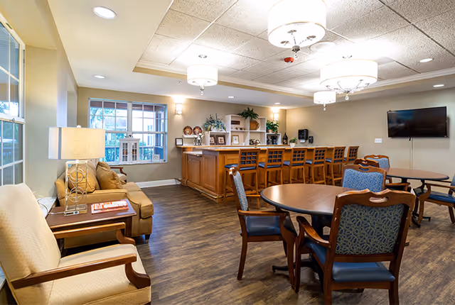 A well-lit common area in a senior living facility featuring a wooden bar with several bar stools, round tables with chairs, a beige couch, an armchair, a side table with a lamp, and a wall-mounted flat-screen TV. The room has wood flooring, a window, and ceiling lights.