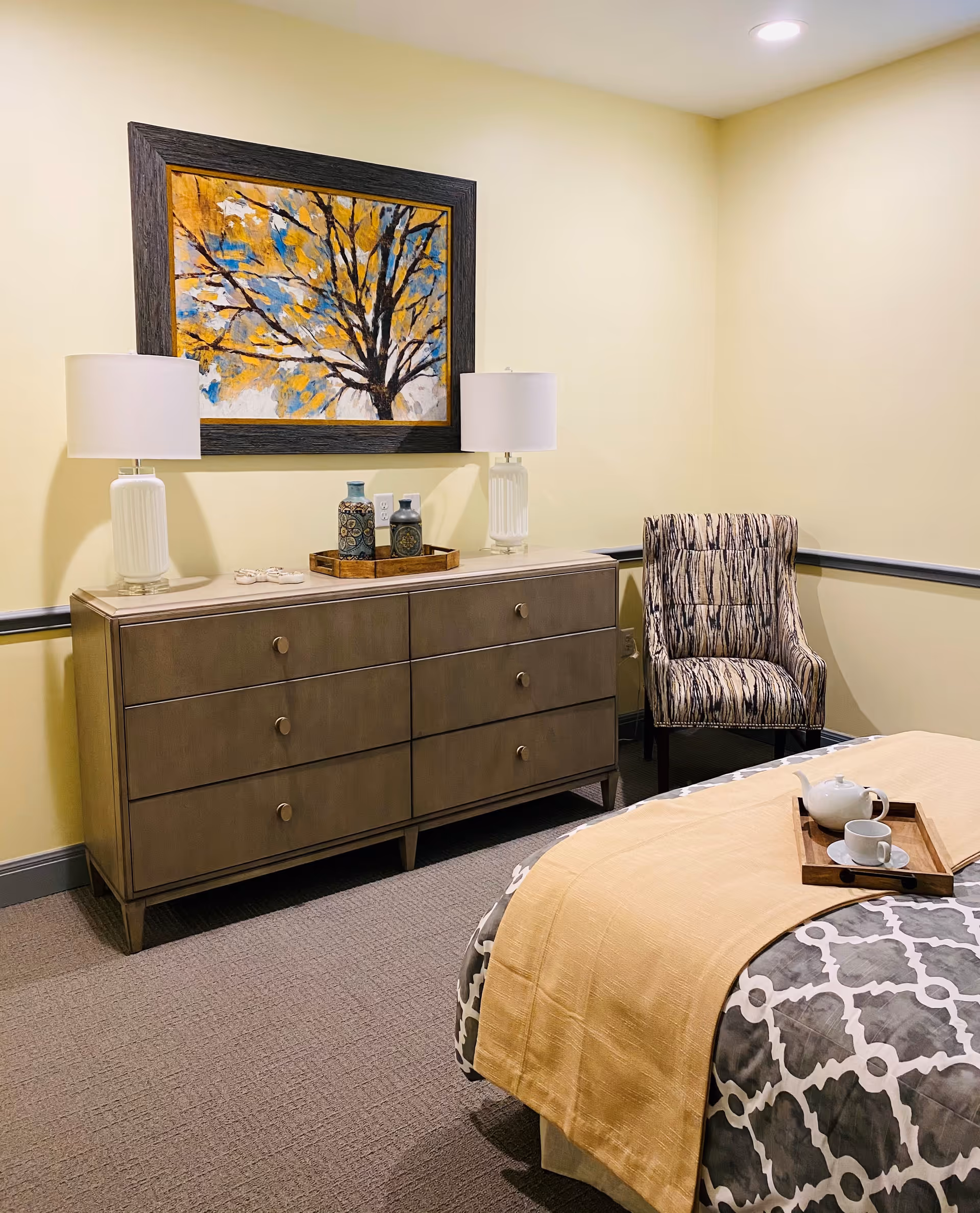 A cozy bedroom corner featuring a wooden dresser with six drawers, two white table lamps, decorative vases on a tray, a patterned armchair, and a bed with a yellow blanket and a tray holding a teapot and cup. A framed painting of a tree with yellow and blue leaves hangs above the dresser.