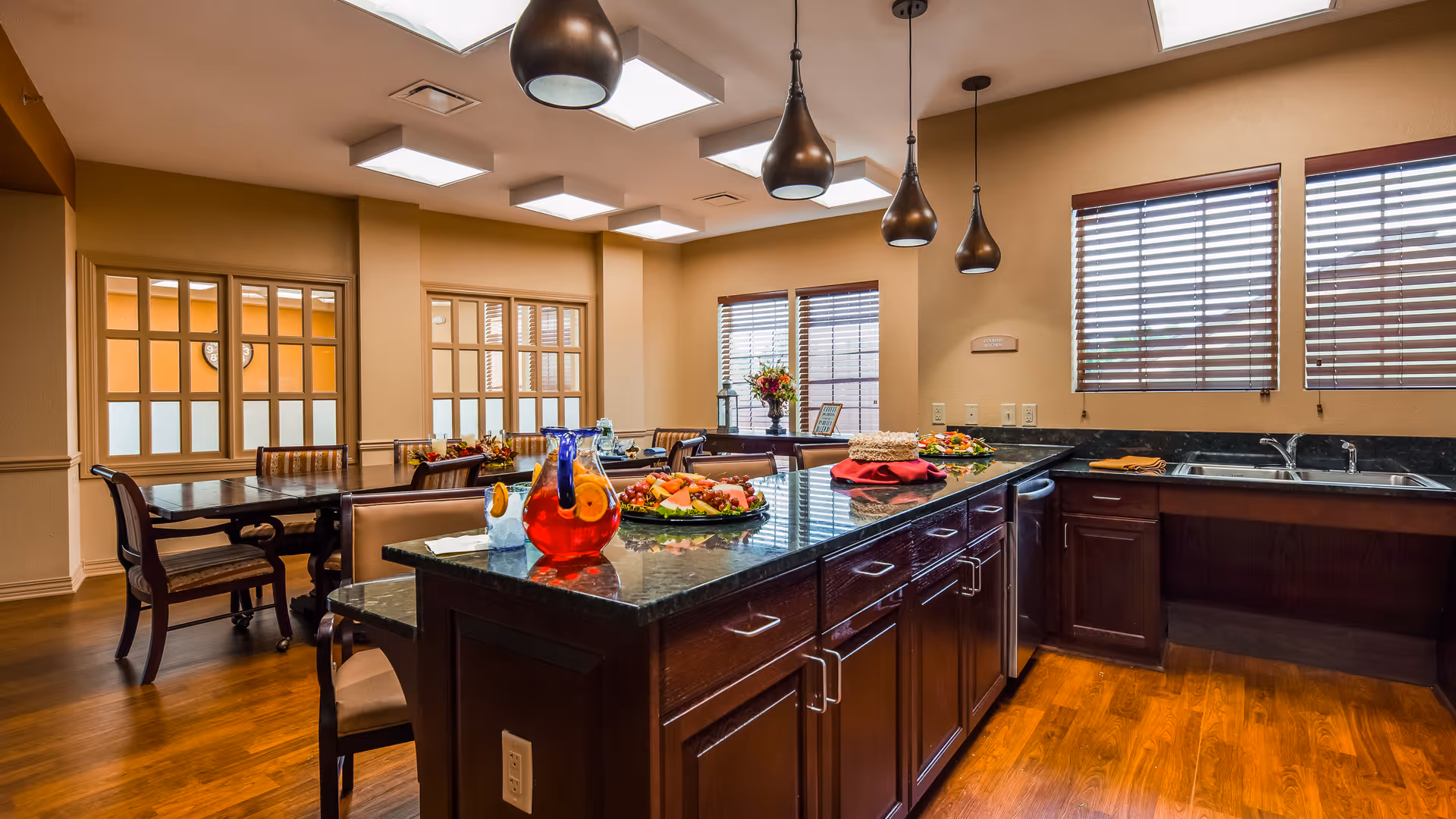 A spacious dining area with a large kitchen island featuring a granite countertop. The island is set with a pitcher of red beverage, a plate of fruit, and other food items. The room has wooden floors, pendant lights hanging from the ceiling, and windows with wooden blinds allowing natural light to enter. In the background, there are dining tables and chairs arranged neatly.