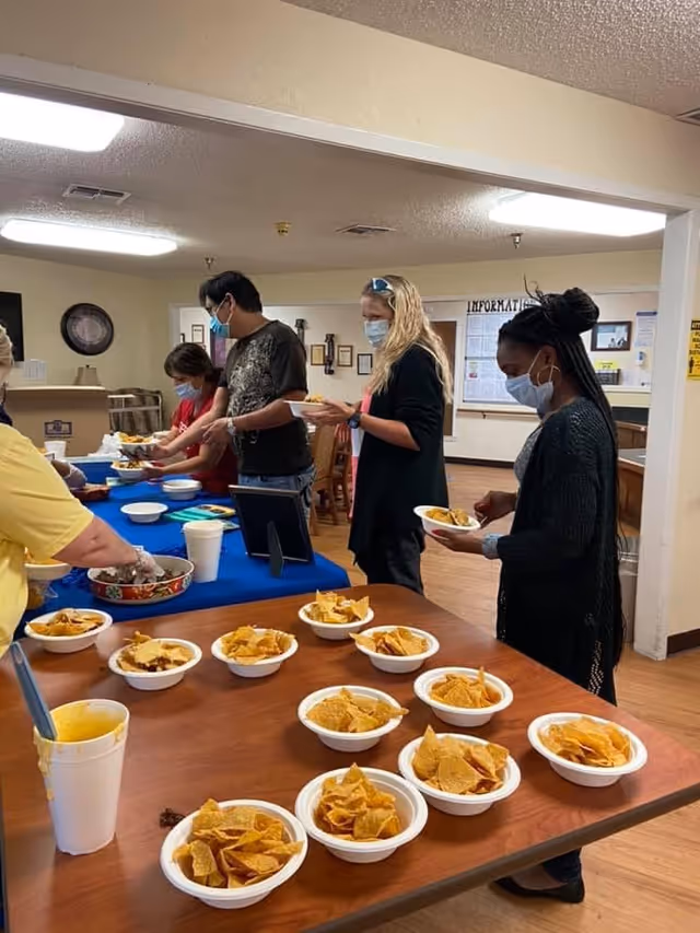 Several people wearing face masks are standing in line serving themselves food from a buffet table inside a room. There are multiple bowls filled with tortilla chips on a wooden table in the foreground. The room has beige walls and fluorescent lighting.