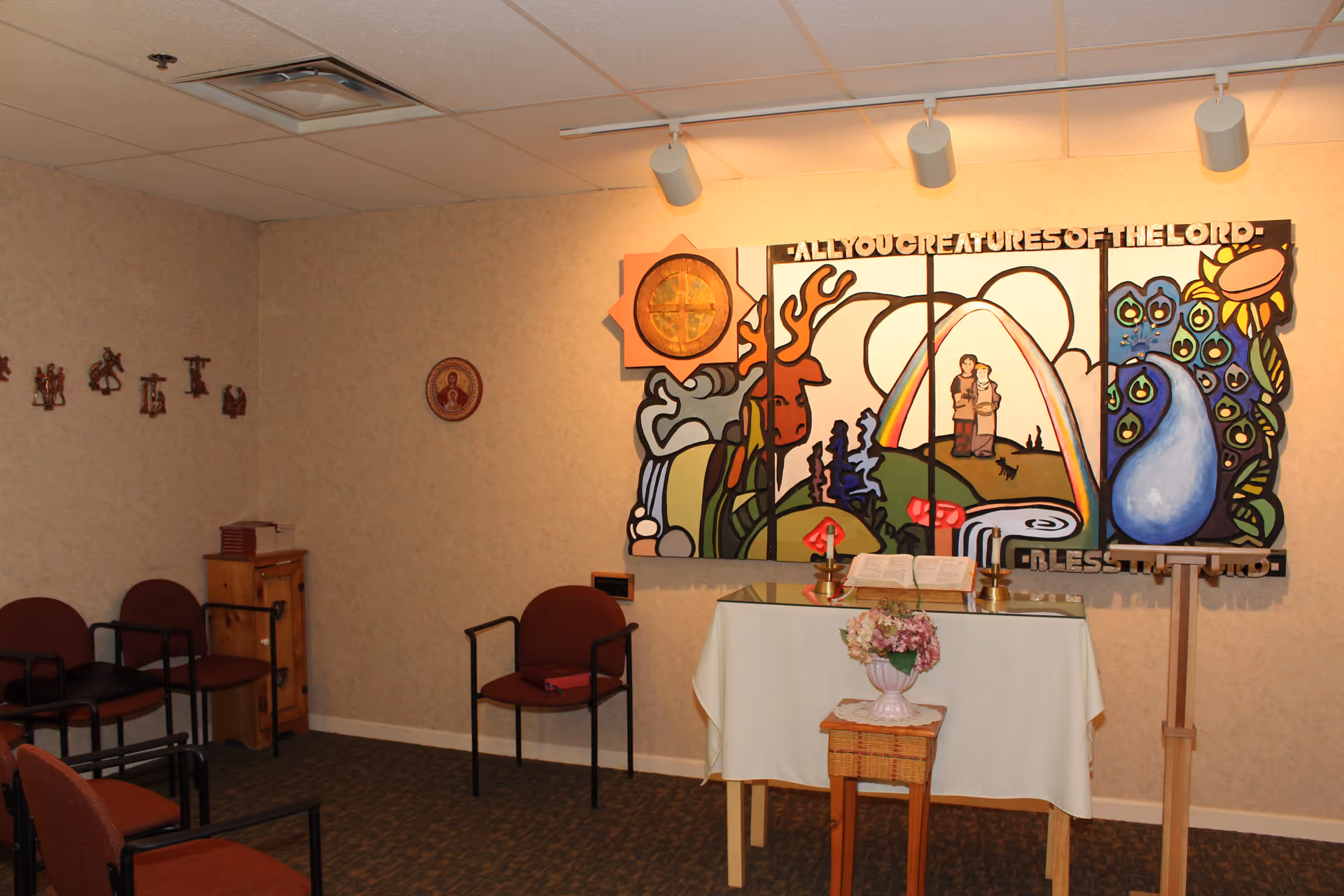 A small chapel room with a table covered by a white cloth holding an open Bible, two candles, and a flower vase on a small stand in front. The wall features a colorful mural with the text 'ALL YOU CREATURES OF THE LORD - BLESS THE LORD'. Several chairs with red cushions are arranged around the room, and religious icons are mounted on the walls.
