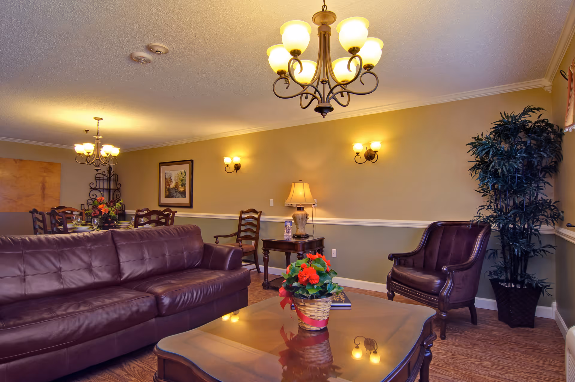 A cozy living room area with a brown leather sofa, a matching armchair, a wooden coffee table with a flower pot, and a side table with a lamp. The room has warm lighting from ceiling chandeliers and wall sconces, with a dining area visible in the background featuring a dining table and chairs. There is a large potted plant in the corner and framed artwork on the wall.