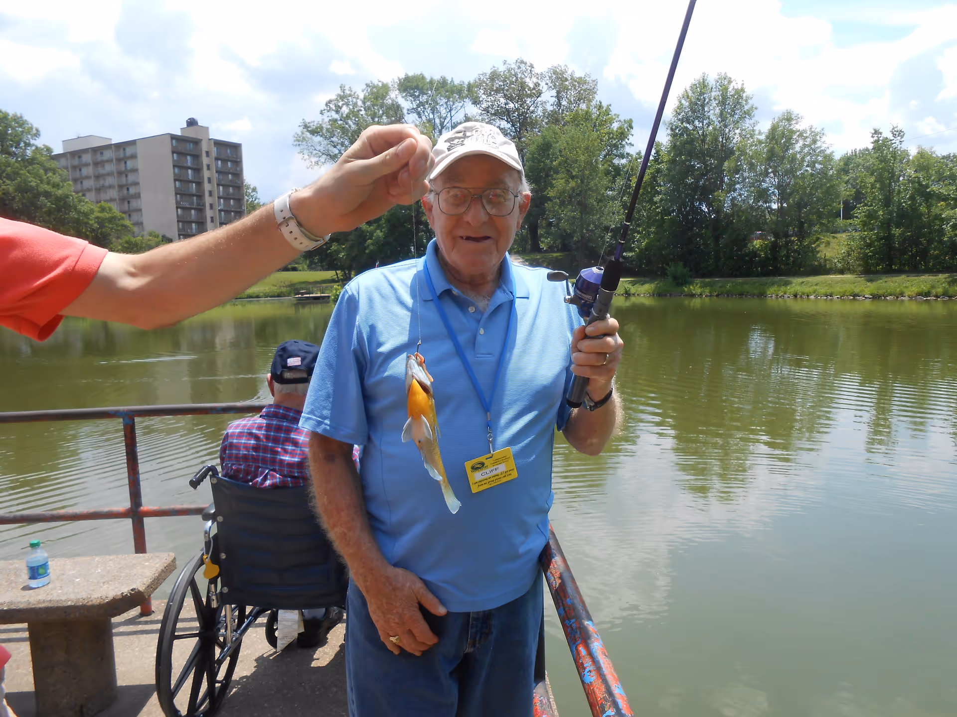 An elderly man wearing a light blue polo shirt, glasses, and a white cap is holding a fishing rod and smiling while standing on a dock by a lake. Another person is holding up a fishing line with a small fish caught on the hook. In the background, there is a man in a wheelchair wearing a plaid shirt and a cap, and trees and a tall building are visible across the lake under a partly cloudy sky.