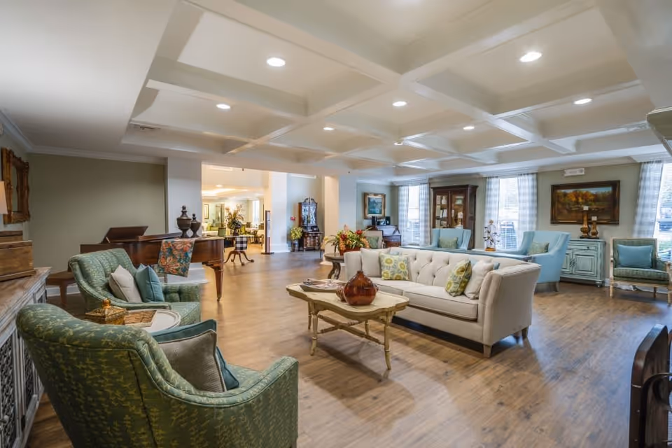 A spacious and well-lit living room area with a beige tufted sofa, green patterned armchairs, a wooden coffee table with decorative items, and large windows with sheer curtains. The room features a coffered ceiling with recessed lighting and wooden flooring. In the background, there is a grand piano and additional seating with blue armchairs and a cabinet.
