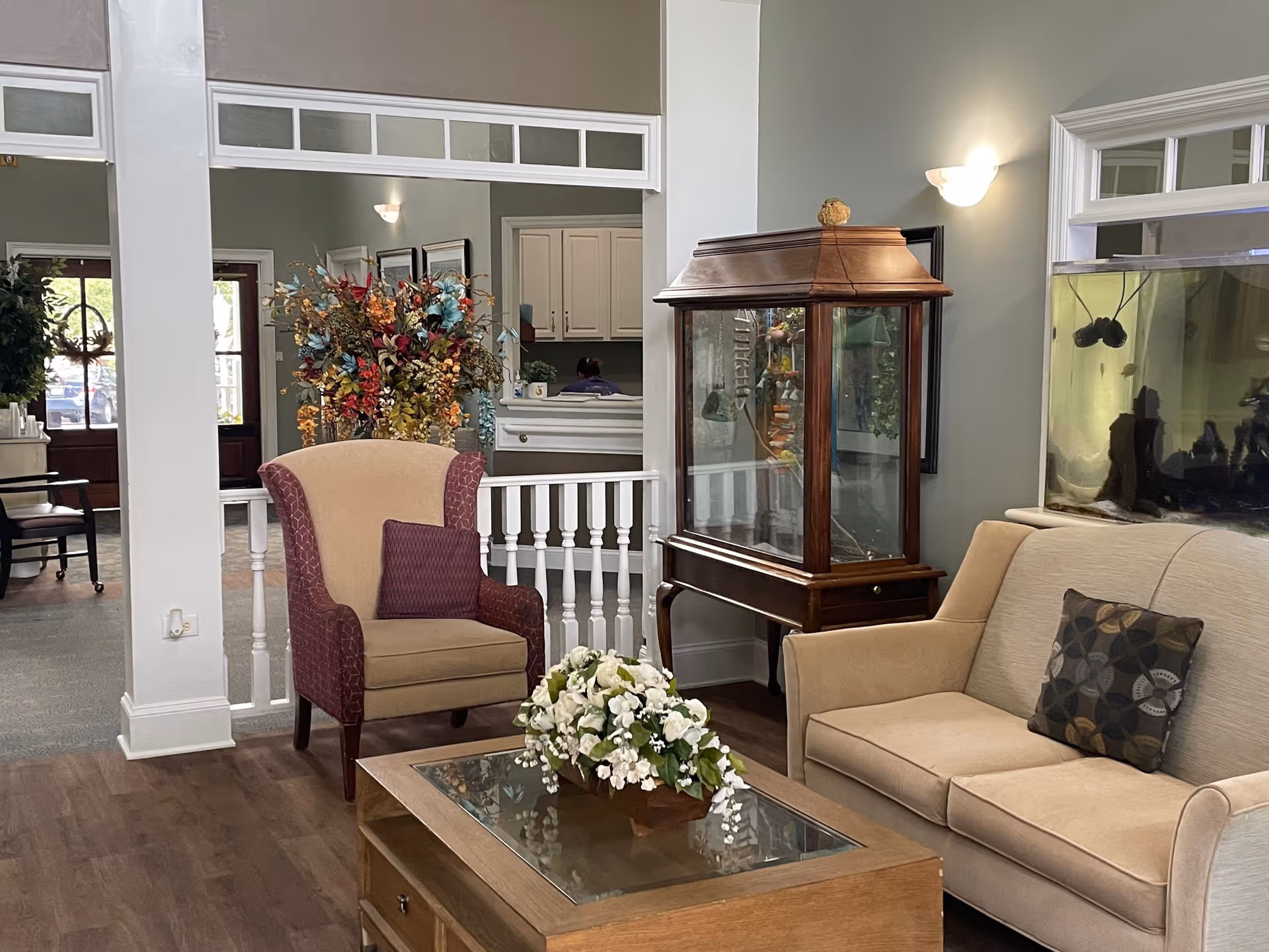 A furnished common area with an armchair and sofa around a coffee table with a floral centerpiece, a wooden display cabinet, and a large fish tank.