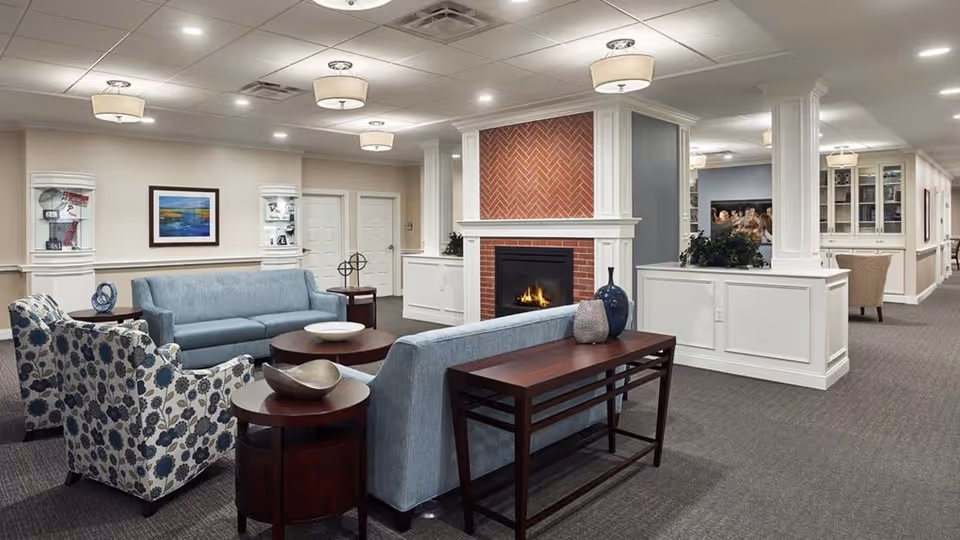 A spacious and well-lit living room area in a senior living facility featuring two light blue sofas, two floral patterned armchairs, a wooden coffee table, and side tables with decorative items. A brick fireplace with a white mantel is centrally located, and the room has recessed lighting and ceiling fixtures. The walls are light-colored with framed artwork and display cabinets.