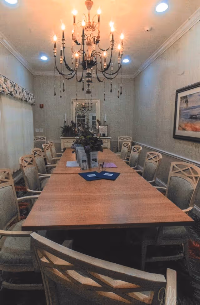 A long wooden conference or dining table surrounded by cushioned chairs with wooden frames in a well-lit room. A large chandelier with multiple lit candles hangs above the table. There is a mirror and decorative items on a sideboard at the far end of the room, and a framed painting on the right wall.