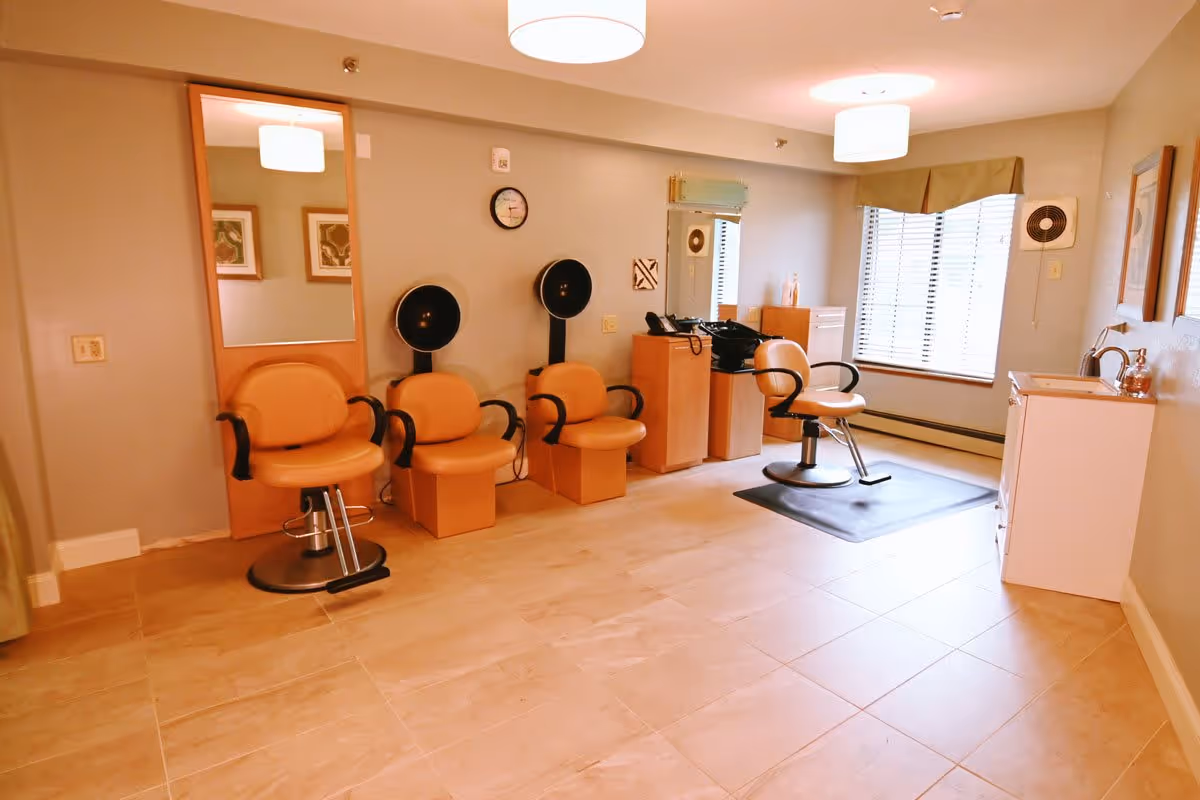 Interior view of a senior living facility hair salon with three orange salon chairs, two hair dryers mounted on the wall, a large mirror, a window with blinds, and a small sink area. The room has beige tiled flooring and soft lighting from ceiling fixtures.