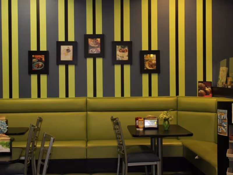 Interior view of a dining area with green cushioned bench seating along the wall, several metal chairs, and black tables. The wall behind the seating has vertical green and gray stripes and is decorated with five framed pictures of food. On the tables are condiments, napkins, and a small vase with flowers.