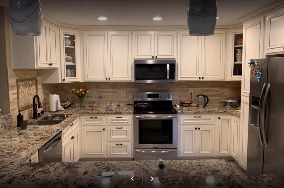 Modern kitchen with white cabinets, granite countertops, stainless steel appliances including a microwave, oven, dishwasher, and refrigerator. The kitchen features a double sink, a vase with orange tulips, a fruit bowl with bananas, and various kitchen items on the counters. Recessed lighting is installed in the ceiling.