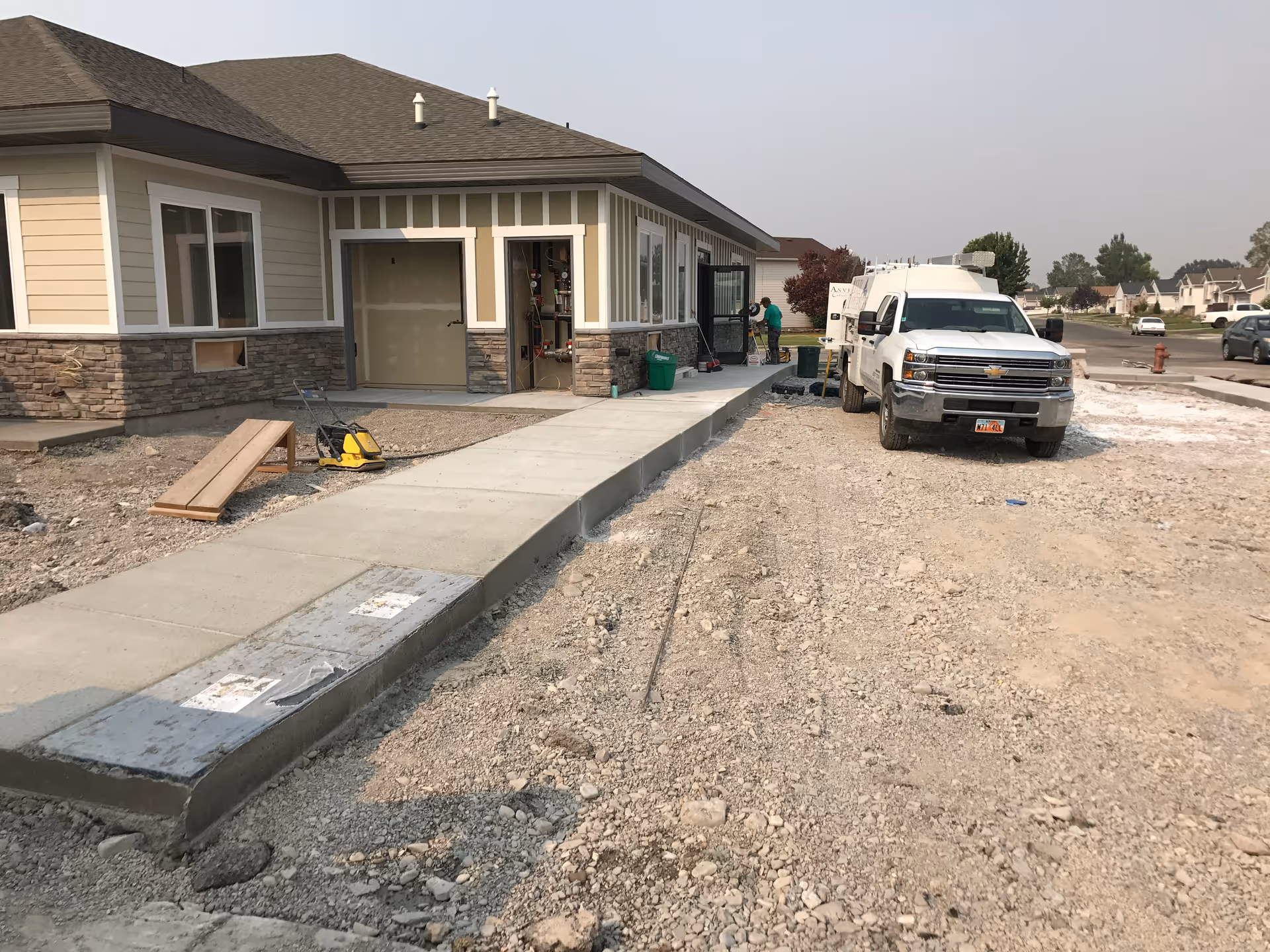 Exterior view of a building under construction with a newly poured concrete sidewalk. A white truck is parked on the gravel area beside the sidewalk, and a person is working near the building entrance. The building has beige siding with stone accents and multiple windows.