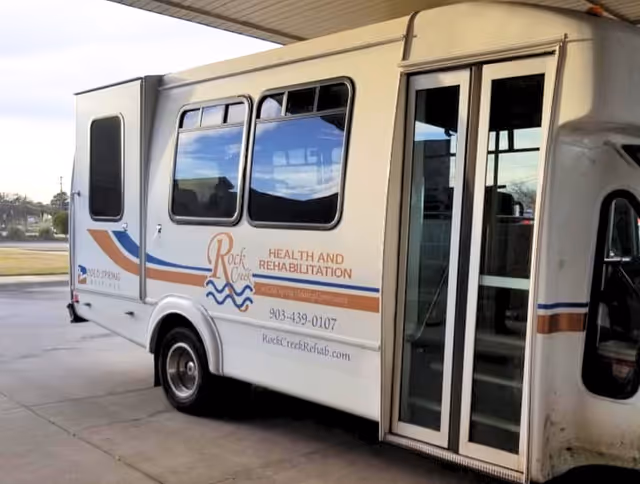 A white shuttle bus parked under a covered area. The bus has signage on the side that reads 'Rock Creek Health and Rehabilitation' along with a phone number and website. The bus door is open, and the background shows an outdoor area with grass and trees.