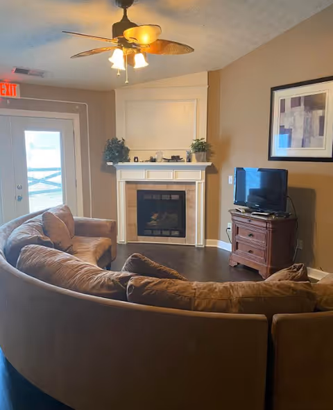 Cozy living room with a curved brown sectional facing a fireplace, TV on a wooden stand, ceiling fan, and a glass exit door.