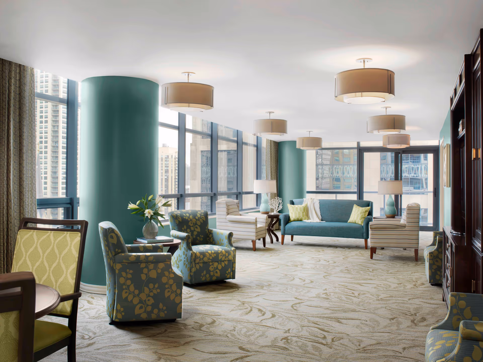 Bright modern communal lounge with a teal sofa, patterned armchairs, large windows showing a city view, and multiple ceiling lamps.