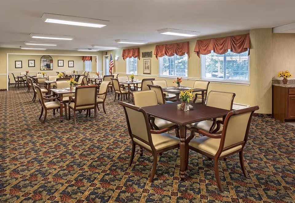 A spacious dining room with multiple tables and chairs arranged neatly. Each table has a small floral centerpiece. The room has patterned carpet flooring, large windows with red valance curtains, and framed artwork on the walls. An American flag is visible near the back wall.