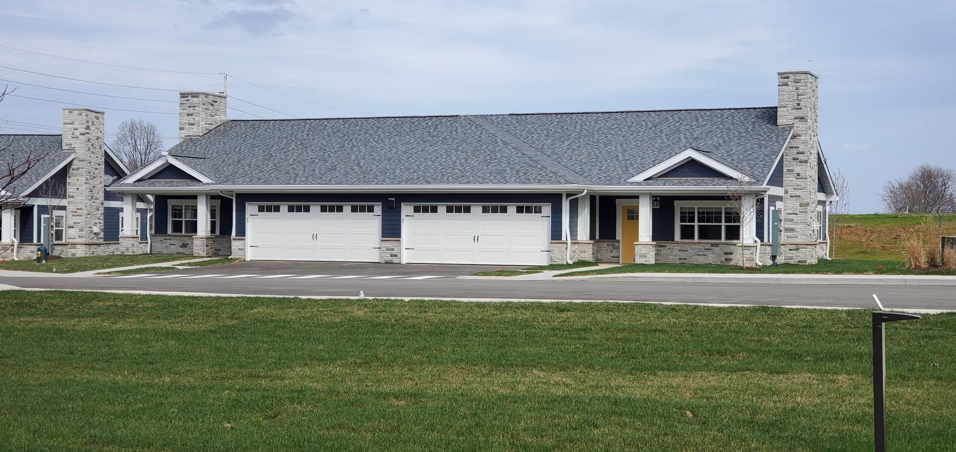 Single-story residential building with three white garage doors, stone chimneys, a covered porch, and a grassy lawn in front.