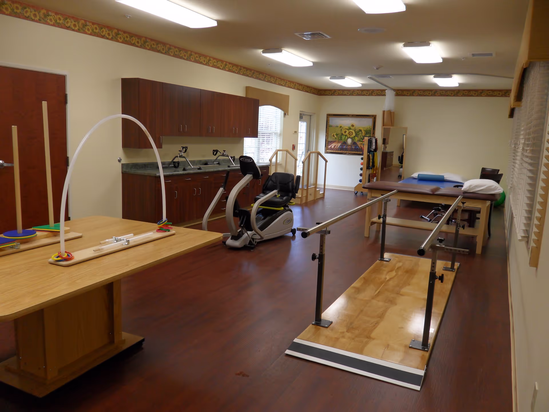 A rehabilitation or physical therapy room with wooden flooring, parallel bars for walking exercises, an exercise bike, therapy tables, and a table with a ring toss game. The room has wooden cabinets along one wall, windows with blinds, and a painting of sunflowers on the far wall.