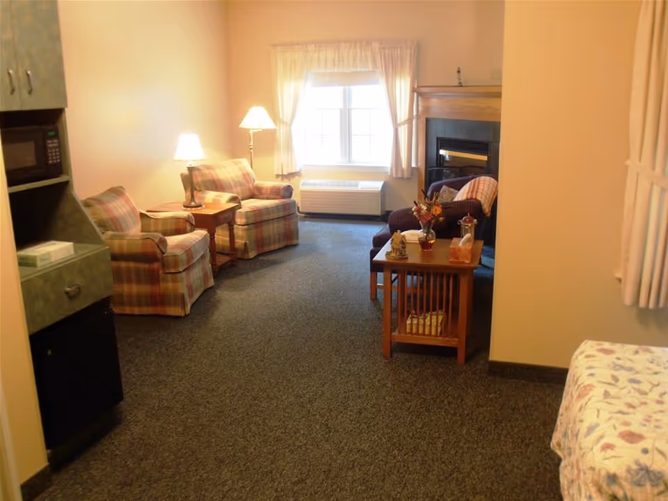 A cozy living room area in a retirement community featuring two plaid armchairs with a small wooden table and lamp between them, a window with white curtains, a fireplace with a dark armchair and a wooden coffee table in front of it, and part of a bed with floral bedding visible on the right side. There is also a small kitchenette area with a microwave and mini fridge on the left.