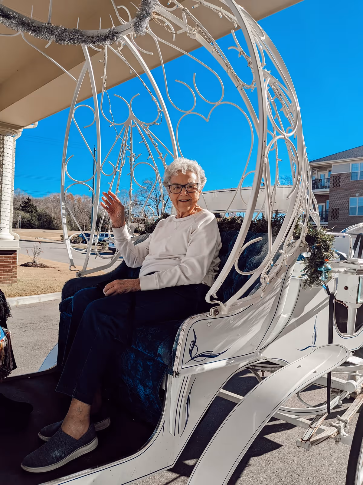 An elderly woman with white hair and glasses sitting and waving while seated in a white decorative horse-drawn carriage outdoors on a sunny day with a clear blue sky.